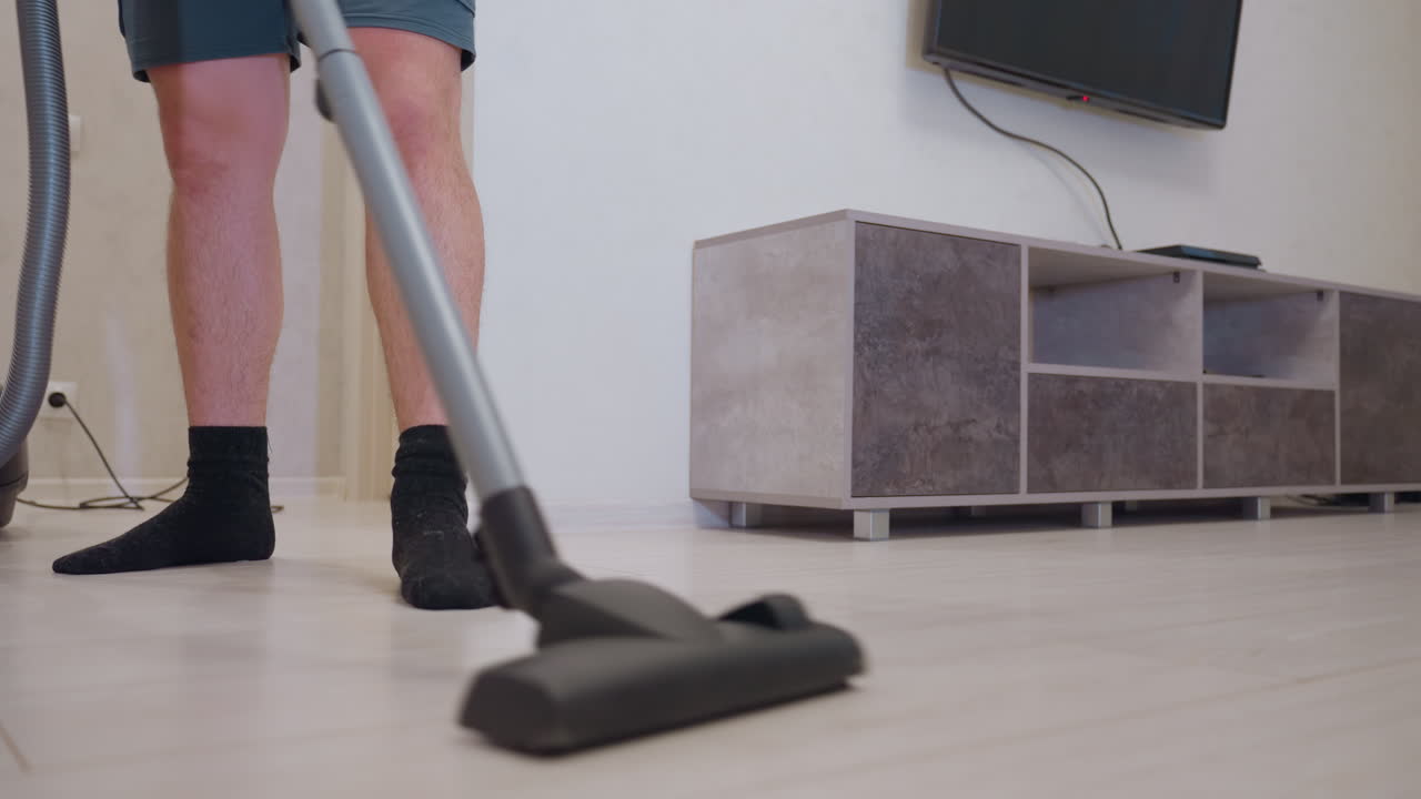 Lower angle view of man in shorts vacuuming light wooden floor while holding hose, standing near modern TV stand and wall mounted television, concept of household cleaning