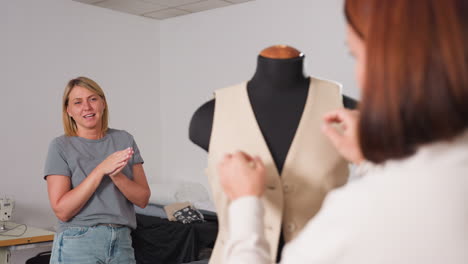 two tailors celebrating successful fitting of beige sleeveless blazer on mannequin in bright workshop, one clapping joyfully while partner adjusts garment fit with precision and shared excitement