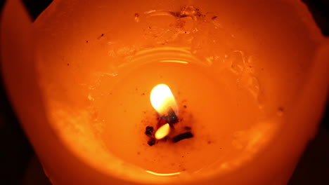 angled close view of an orange and red candle with a litted shaky flame and melted wax on dark background