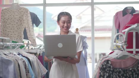 asian female owner of clothing store enjoys using a laptop going on social media and making online marketing after check stock on rails