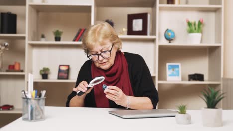 old woman sitting at the table examines the desk clock through a magnifying glass