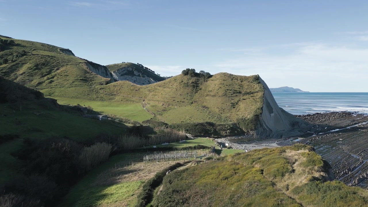 Veduta aerea che sorvola una valle con colline verdi, rivelando la splendida spiaggia di Sakoneta e le sue uniche formazioni rocciose di flysch in una giornata di sole nei Paesi Baschi, Spagna.