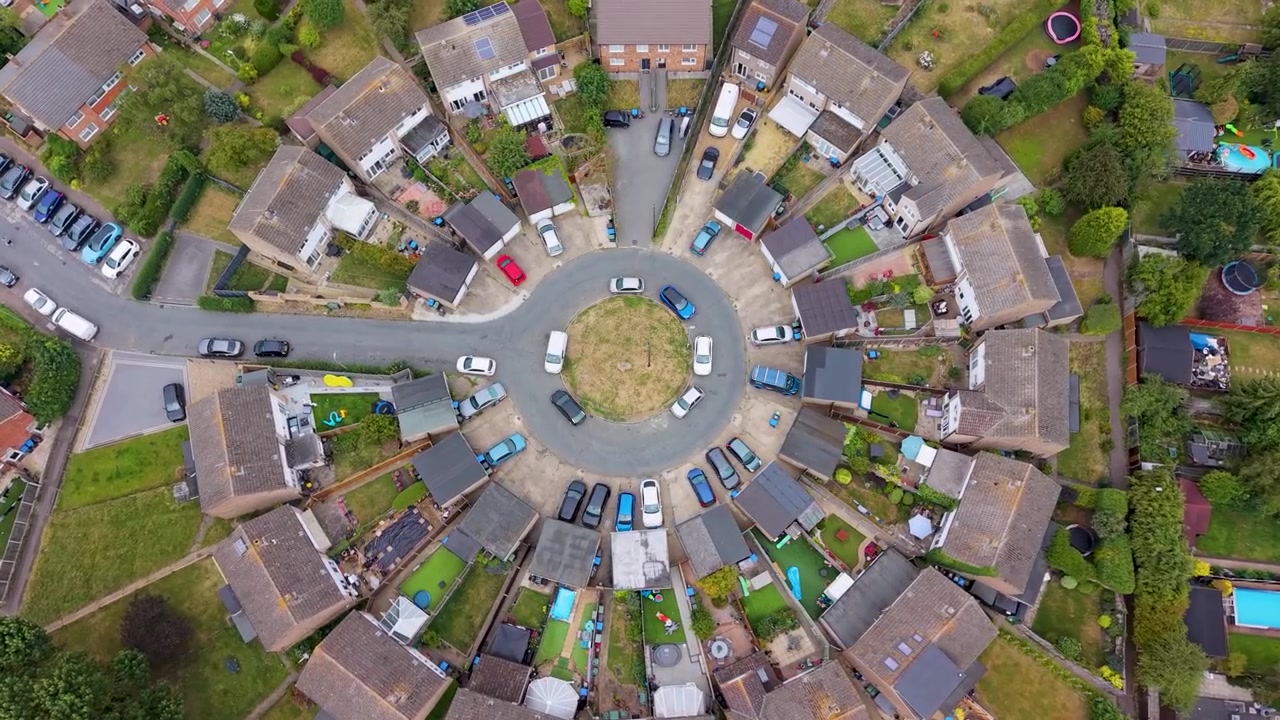 Tomada de drone rotativa descendente sobre Roundhills, um condomínio residencial circular sem saída em Waltham Abbey, Essex, revelando telhados radiais, jardins e carros estacionados abaixo
