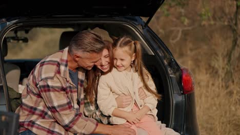 A-happy-middle-aged-brunette-man-with-gray-hair-in-a-checkered-brown-shirt-hugs-his-wife-and-little-daughter-in-the-open-trunk-of-a-black-car-during-his-summer-picnic-outside-the-city-in-the-evening