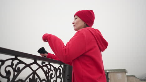 vista lateral de primer plano de una mujer con capucha roja y gorra de pie cerca de una barandilla decorativa de metal, mirando pensativa a la distancia brumosa, ambiente invernal sereno con iluminación suave y fondo borroso