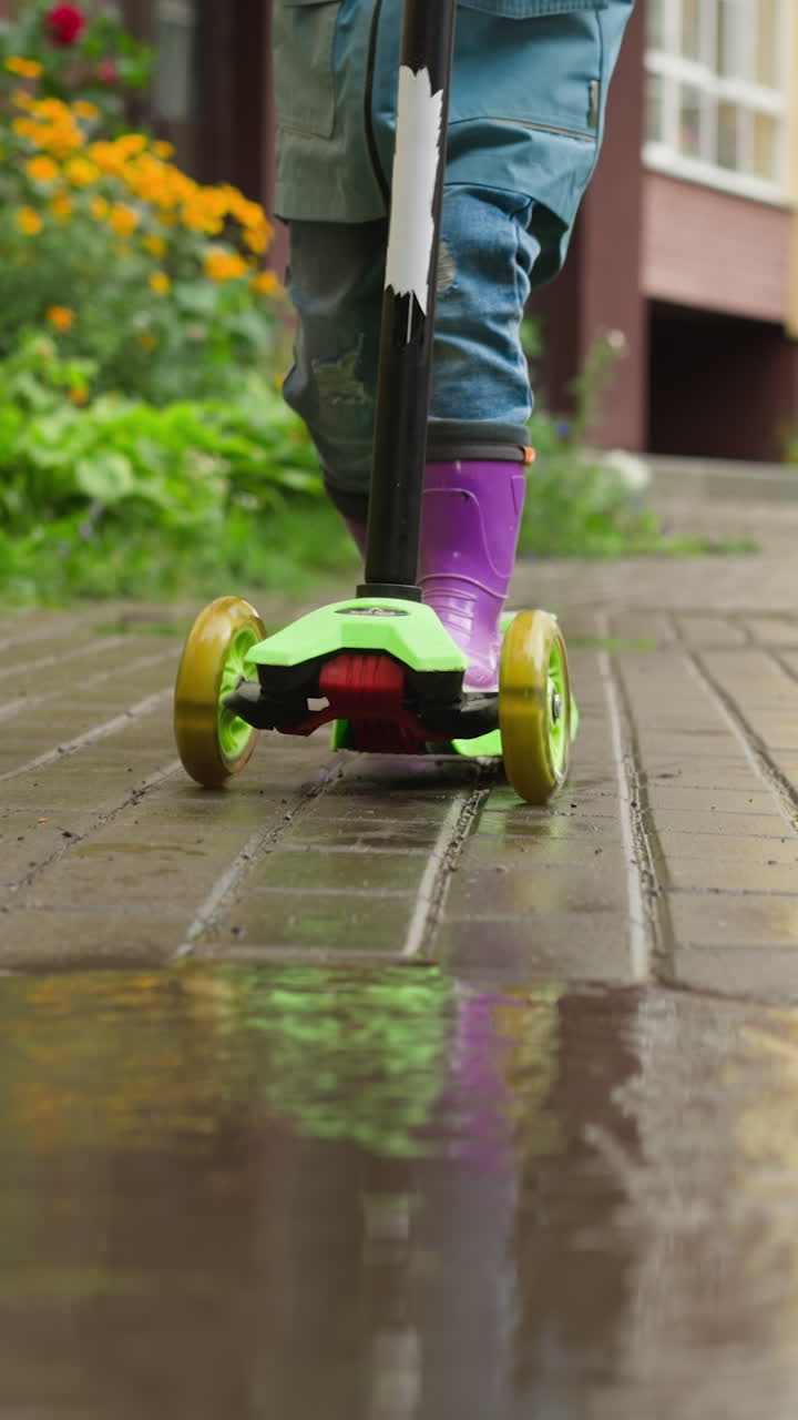 niño corre a través de las gotas de lluvia en scooter. niño con botas de goma se detiene frente al charco en el pavimento. scooter de tres ruedas talla el camino de la alegría a través de la lluvia