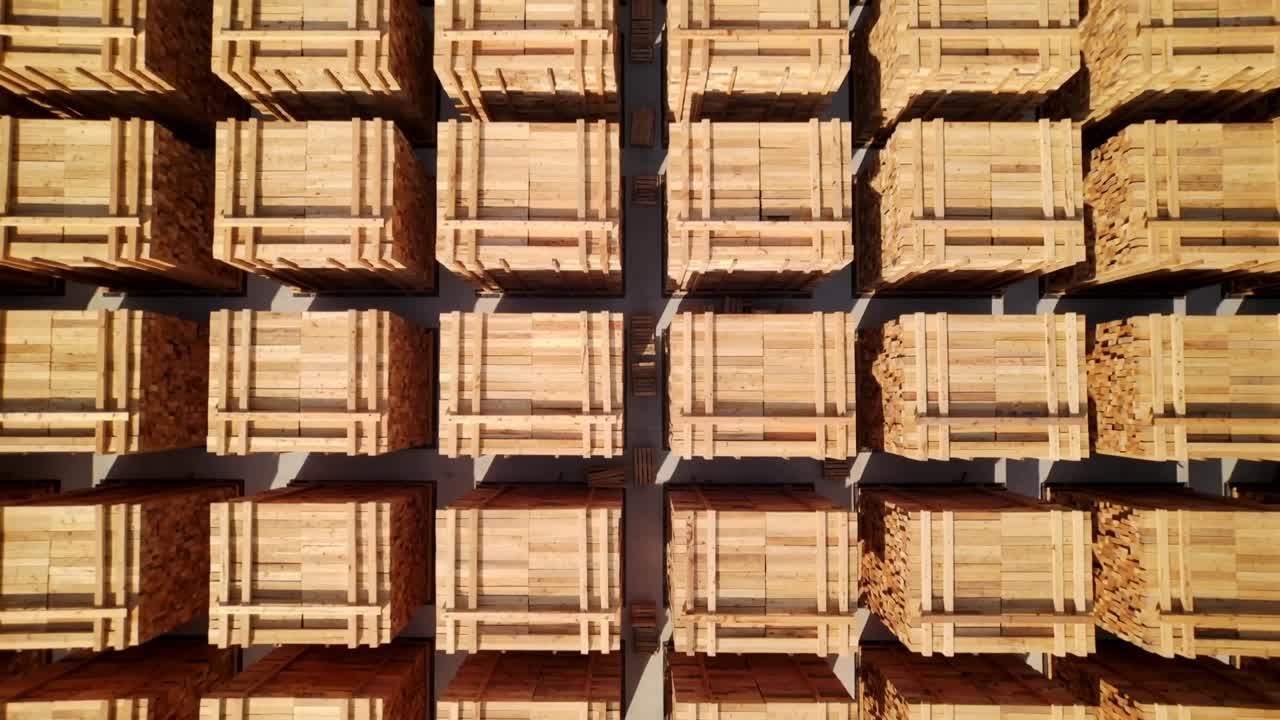 Dronekamera trækker sig tilbage og afslører et tømmerlager med pallekasser, bundtet tømmer og betonstier.
