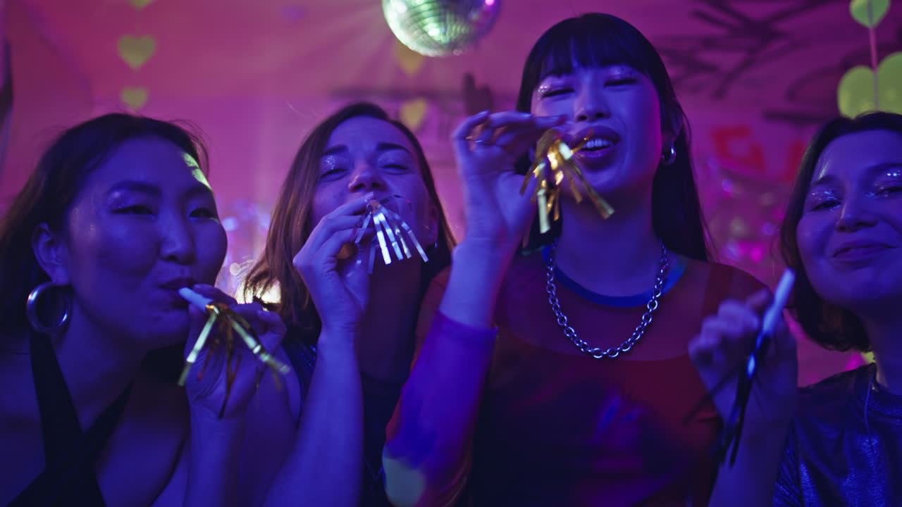 Smiling women blowing noisemakers in neon-lit festivity closeup. Happy girls