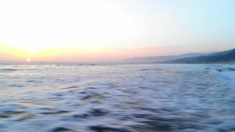 Aerial-shot-flying-low-and-fast-over-the-ocean-waves-crashing-on-the-beach-under-a-colorful-sunset-sky-background-along-the-coast-of-Ventura,-California-DRONE