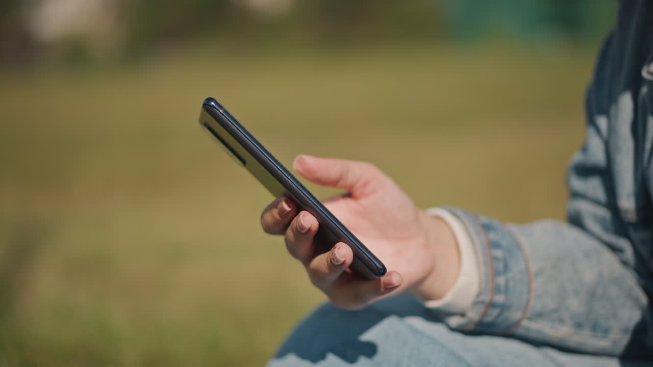 Close-up de mão segurando telefone ao ar livre, manga jeans visível sobre parque gramado e luz solar suave, gesto de rolagem lenta na tela sensível ao toque transmite navegação e mensagens relaxadas, profundidade de campo rasa sutil, caloroso.