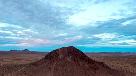 a volcanic cone-shaped mountain or butte in the mojave desert with colorful clouds over the barren and rugged geology - aerial stationary time lapse