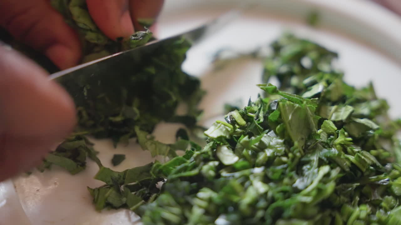 Primo piano di mani che tritano erbe fresche su un piatto bianco. Un cuoco casalingo prepara verdure a foglia in pezzi fini per guarnire l'insalata. Luce naturale calda, focus tattile sul coltello e sulla lama, in una cucina aromatica.