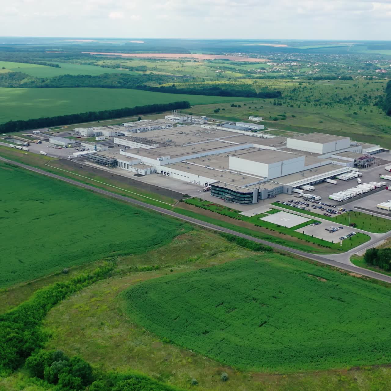 Large industrial zone on green field. Factory surrounded by beautiful landscape in summer. Aerial view. Camera moving back