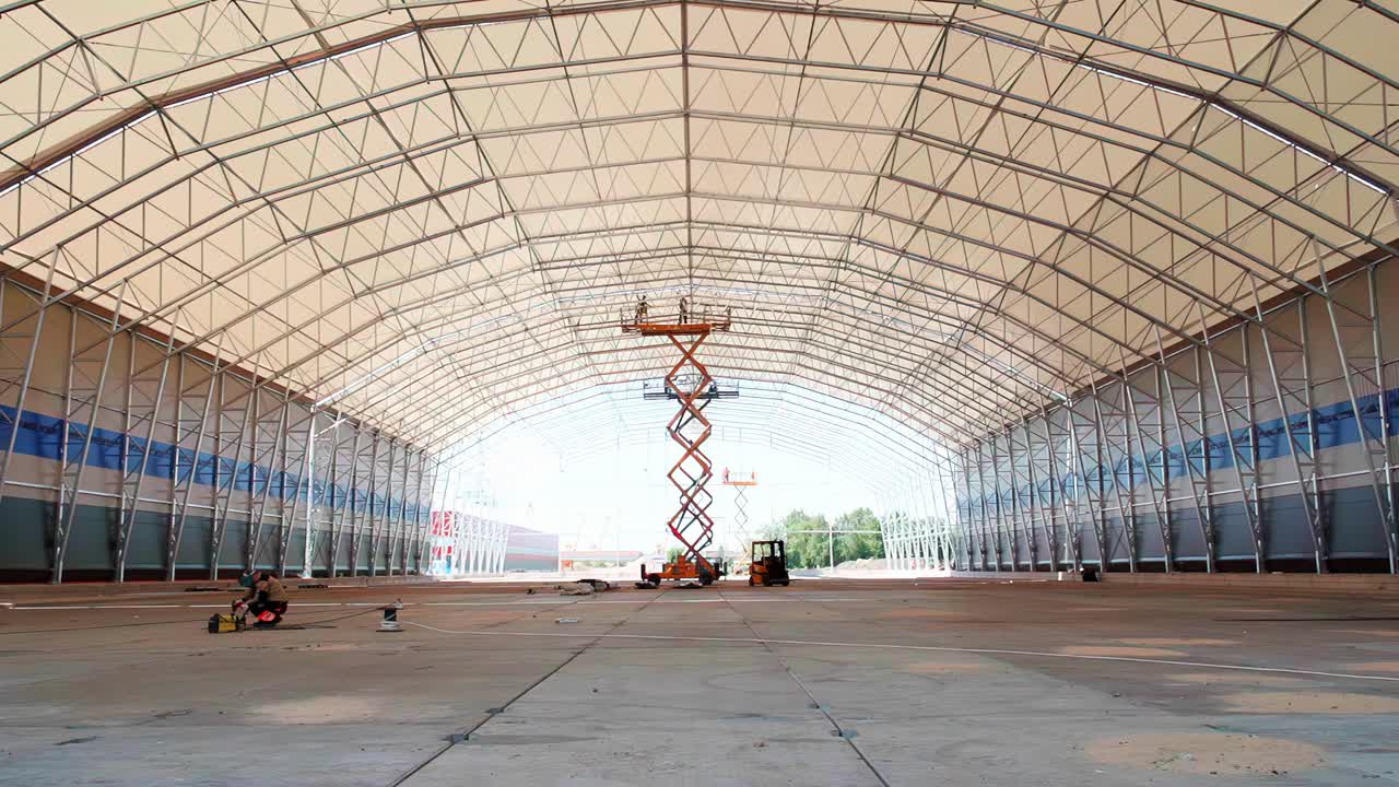 A vast, wide-angle interior view of a massive modern hangar or warehouse under construction, showing the steel truss roof structure and a scissor lift on the concrete floor