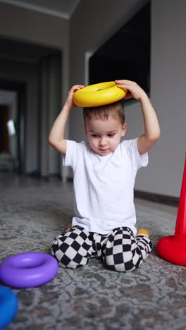 dark-haired baby boy puts the piece of toy pyramid on his head. a toy drops on the floor and kid cheers happily.
