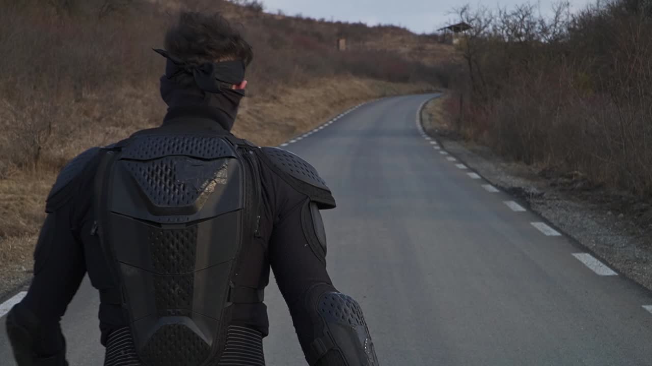 Cinematic back view of a lone man in dark gear holding guns, walking on a winding road. Action vibe, perfect for thriller scenes or storytelling visuals