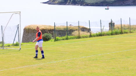 male goalkeeper wearing orange vest catching football and kicking out on pitch, copy space