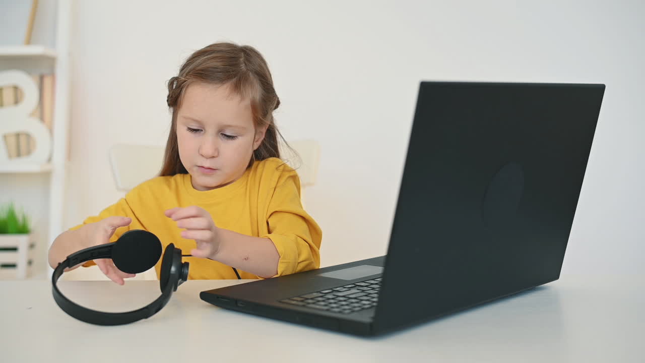 헤드폰을 끼고 있는 어린 소녀는 집에서 노트북을 사용합니다