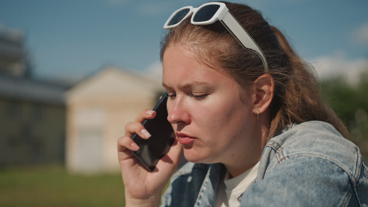 Caucasian woman outdoors making phone call, denim jacket, white sunglasses perched on head, concerned expression, holding mobile to ear, sunlit suburban houses in background, candid closeup, natural
