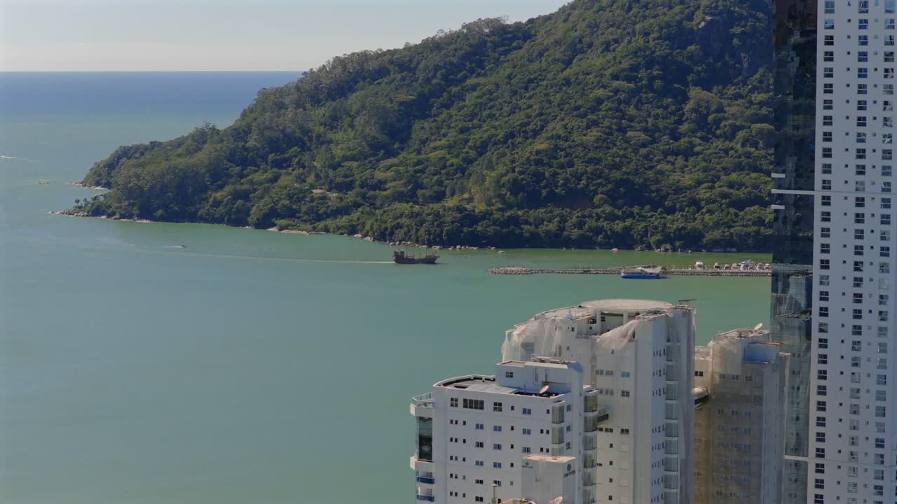 Drone advances over Barra Sul’s mix of city buildings, ocean, boat wake, and green hills