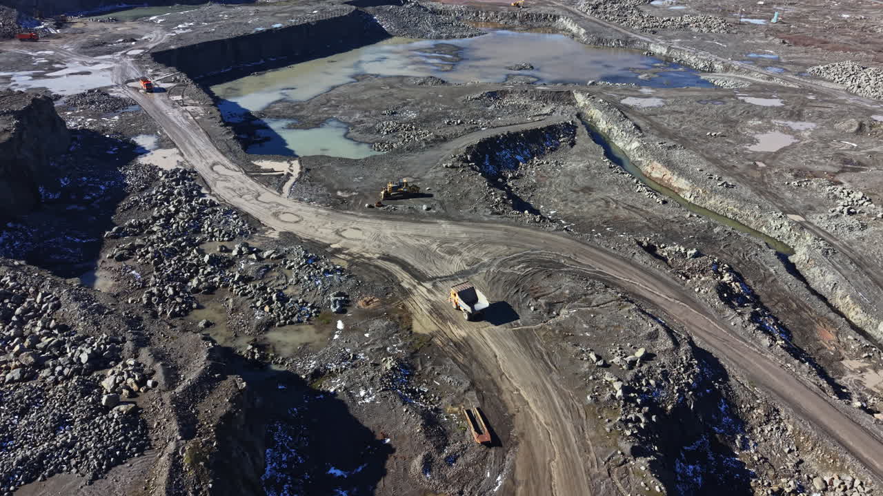 Workers operate machinery in a granite quarry during daylight near a water body and rocky terrain with piles of granite
