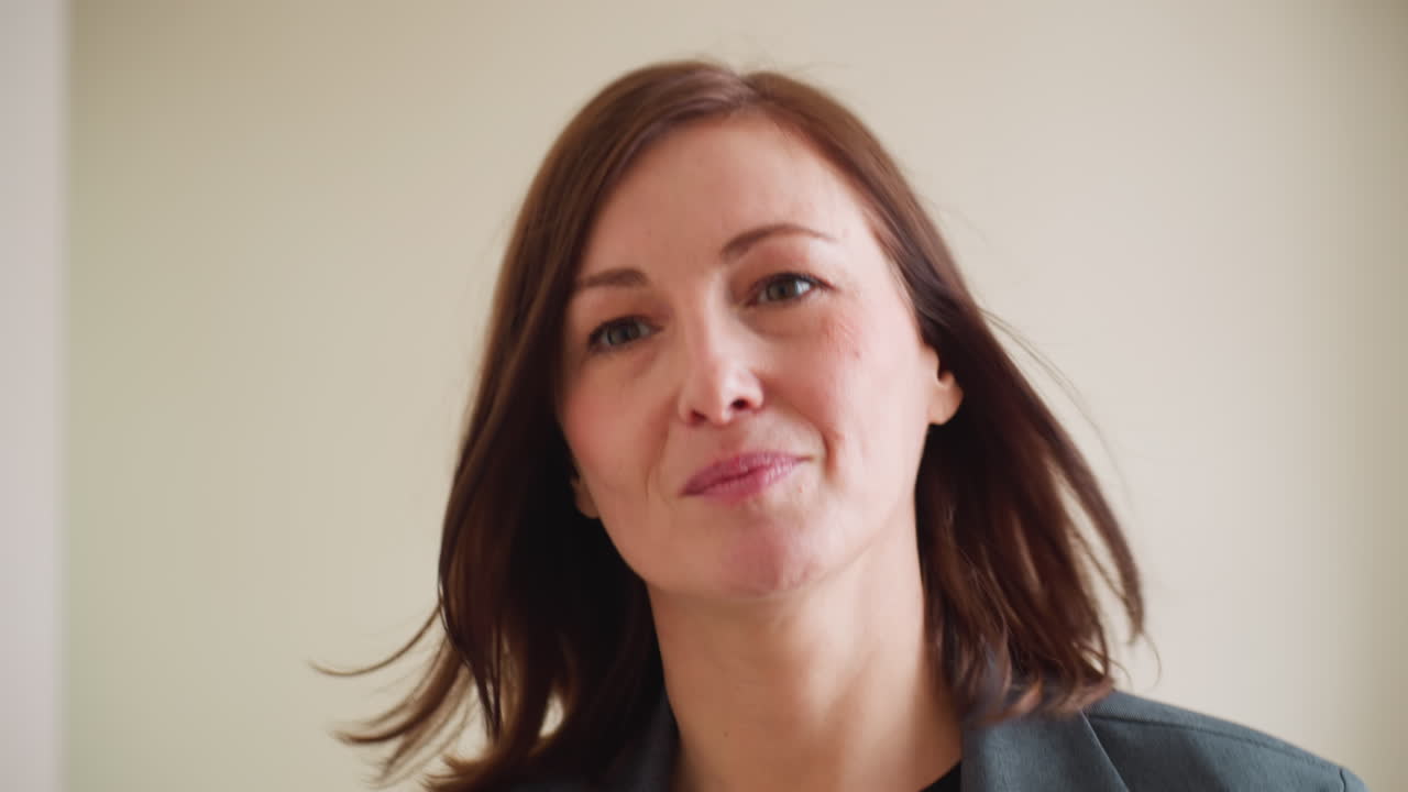 Closeup portrait of confident businesswoman with light makeup and natural smile looking forward with calm determination, hair gently moving, dressed in formal suit against plain office background