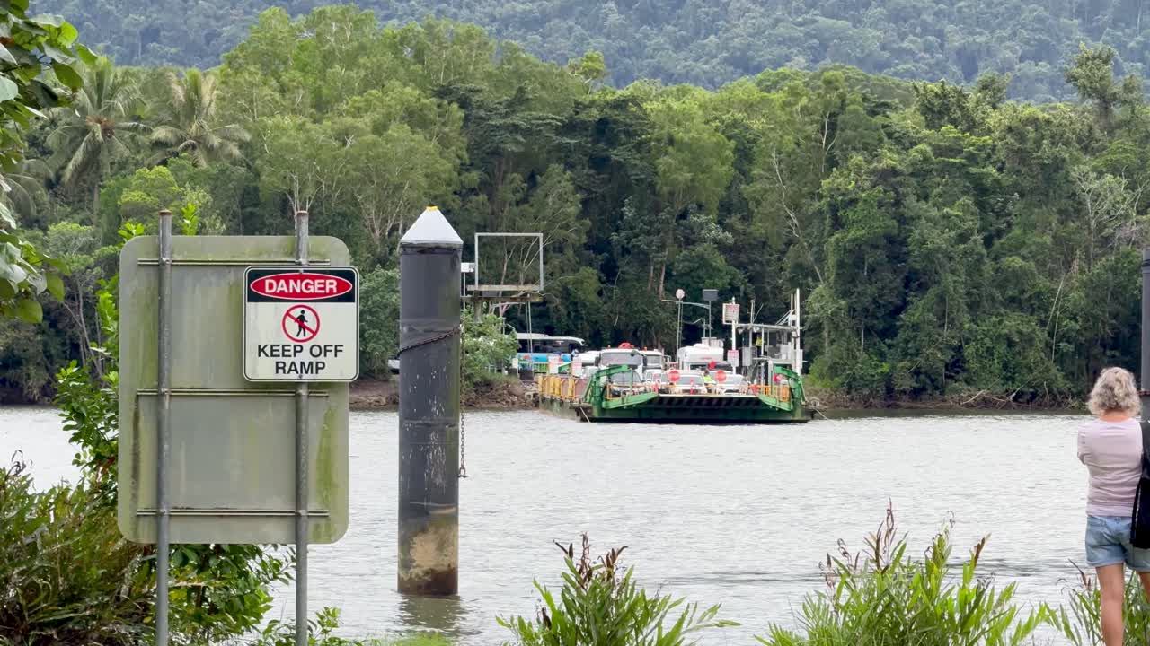 Vehicle ferry nears riverbank, lush rainforest background, daylight, static camera, person observing