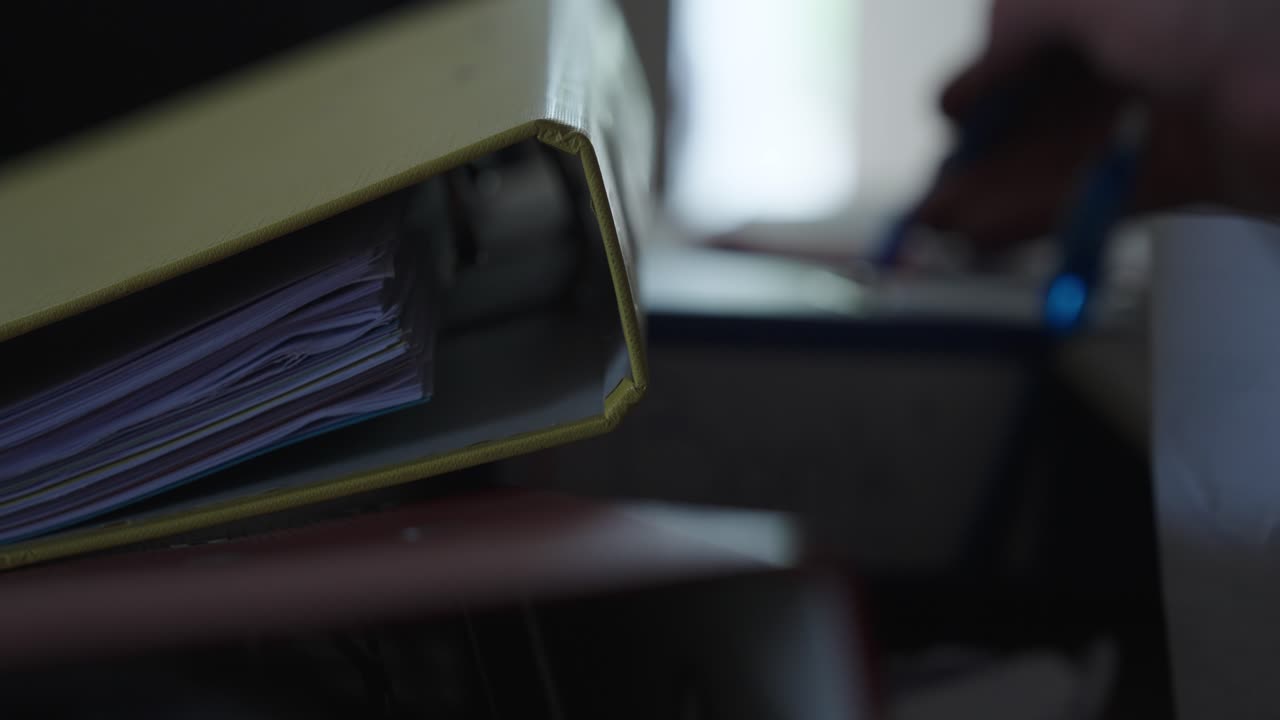 A yellow binder dominates the foreground while someone writes in the background—capturing a quiet moment of focused work or documentation
