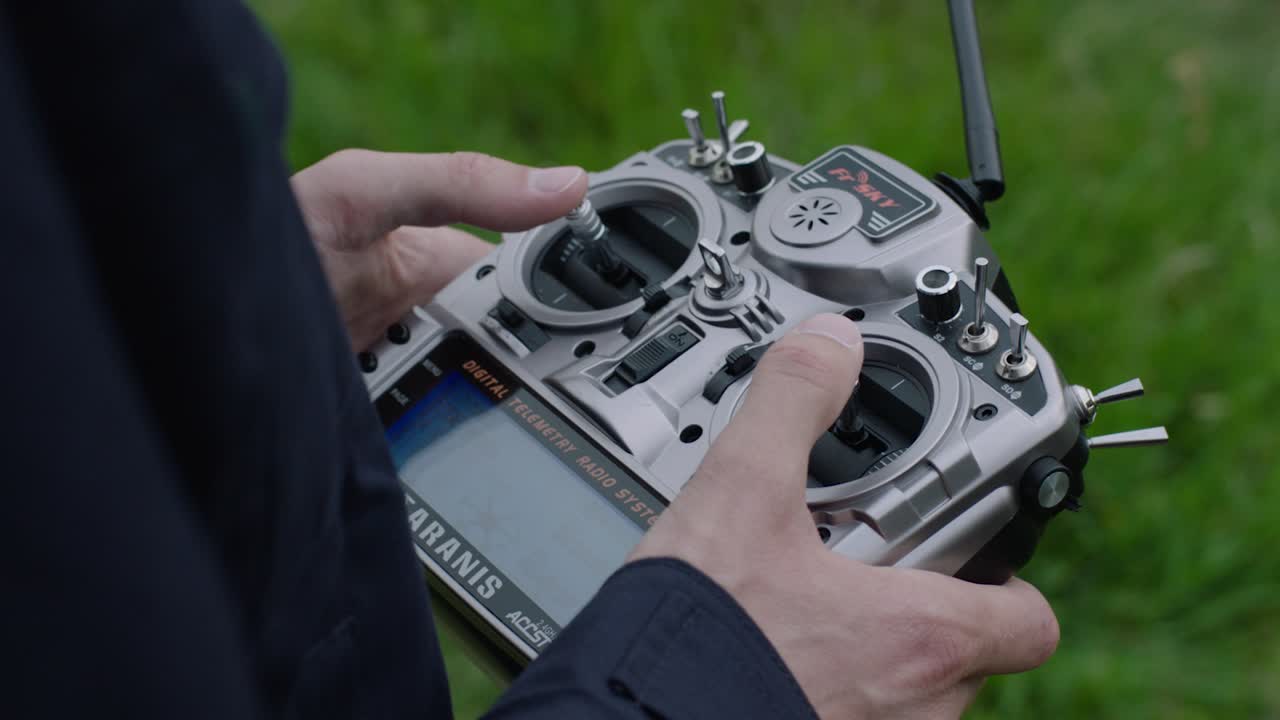 Handheld camera focuses on person's hands holding a silver-black drone remote control, thumbs on joysticks, against a blurred field backdrop