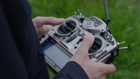 Handheld camera focuses on person&#039;s hands holding a silver-black drone remote control, thumbs on joysticks, against a blurred field backdrop