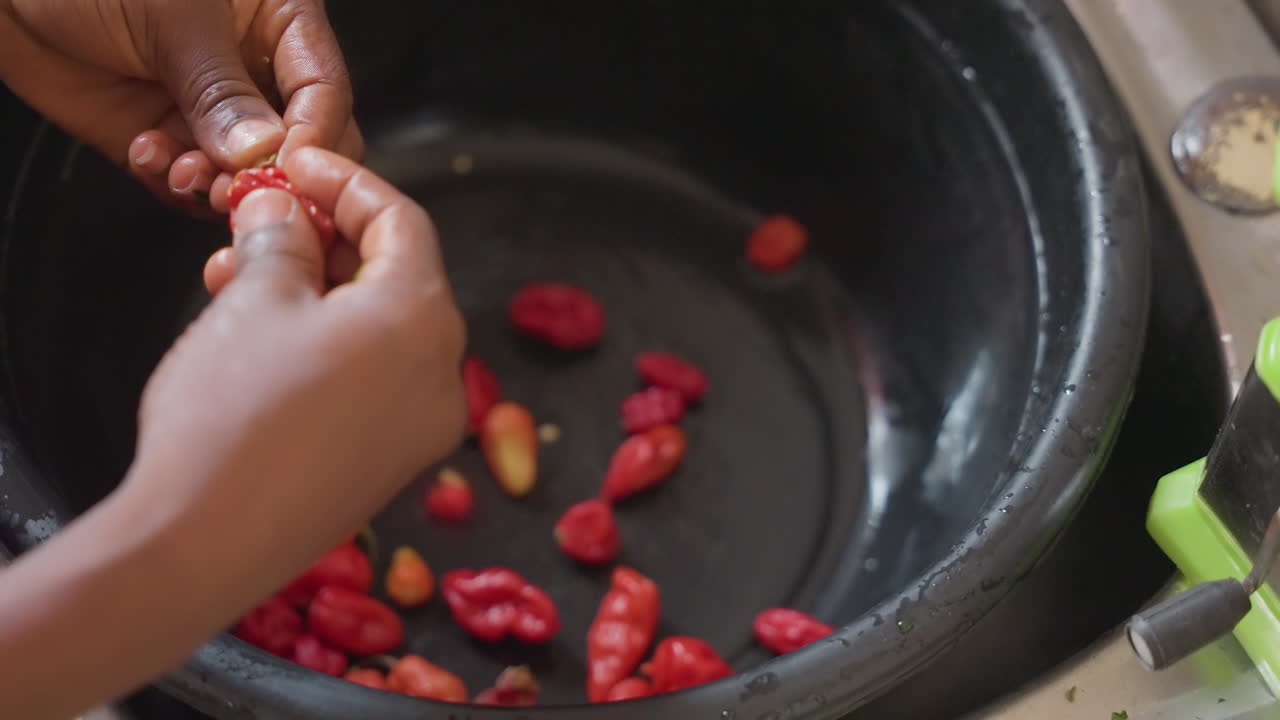 Hands sorting red chilies in basin, home cook carefully inspects and selects small peppers in black basin at sink, closeup tactile sequence shows wet skins, finger movements, vibrant color and calm