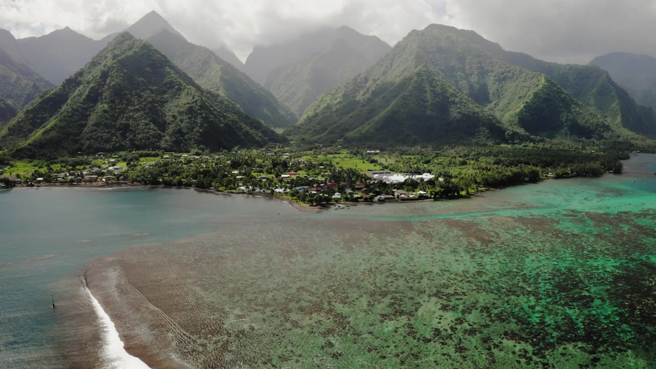 Dronekuva Teahupoon kylästä, Tahitilla, joka sijaitsee sademetsävuorten ja koralliriutan välissä, kattoja hajallaan rannikolla