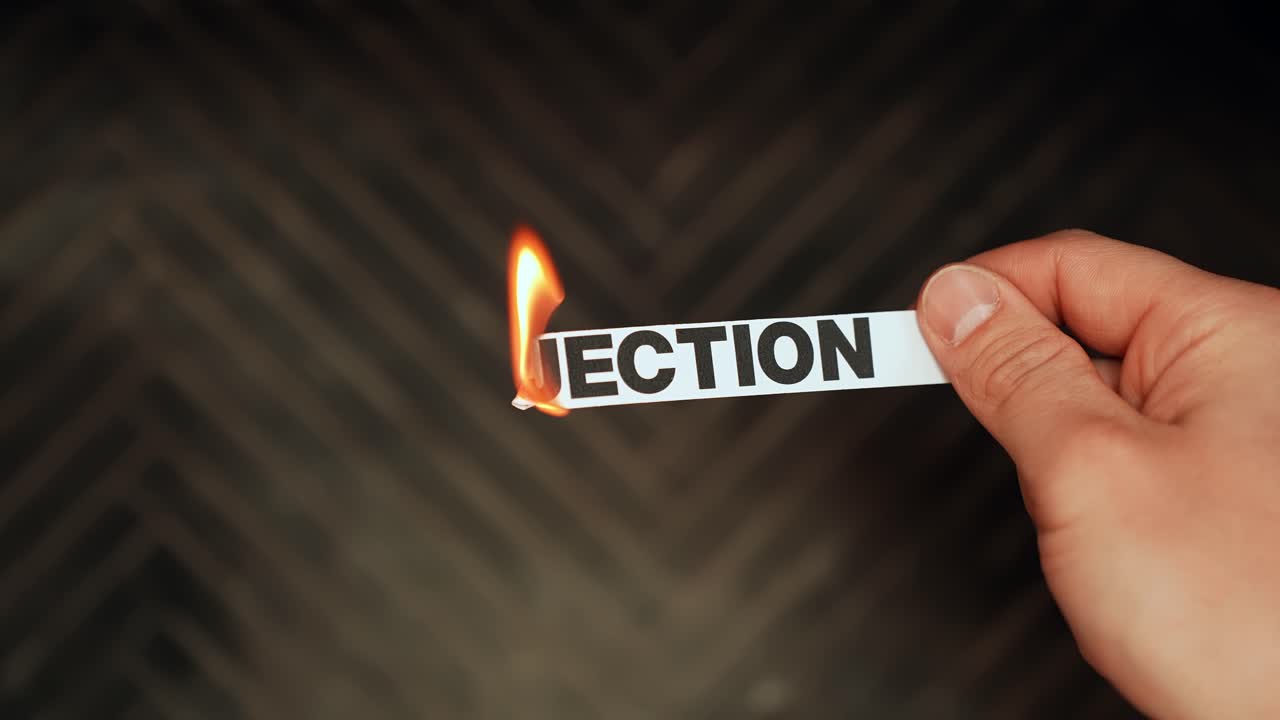 Close-up of a hand holding a paper strip reading “REJECTION” as the edge burns with a bright flame against a dark blurred background, symbolizing emotional pain and letting go