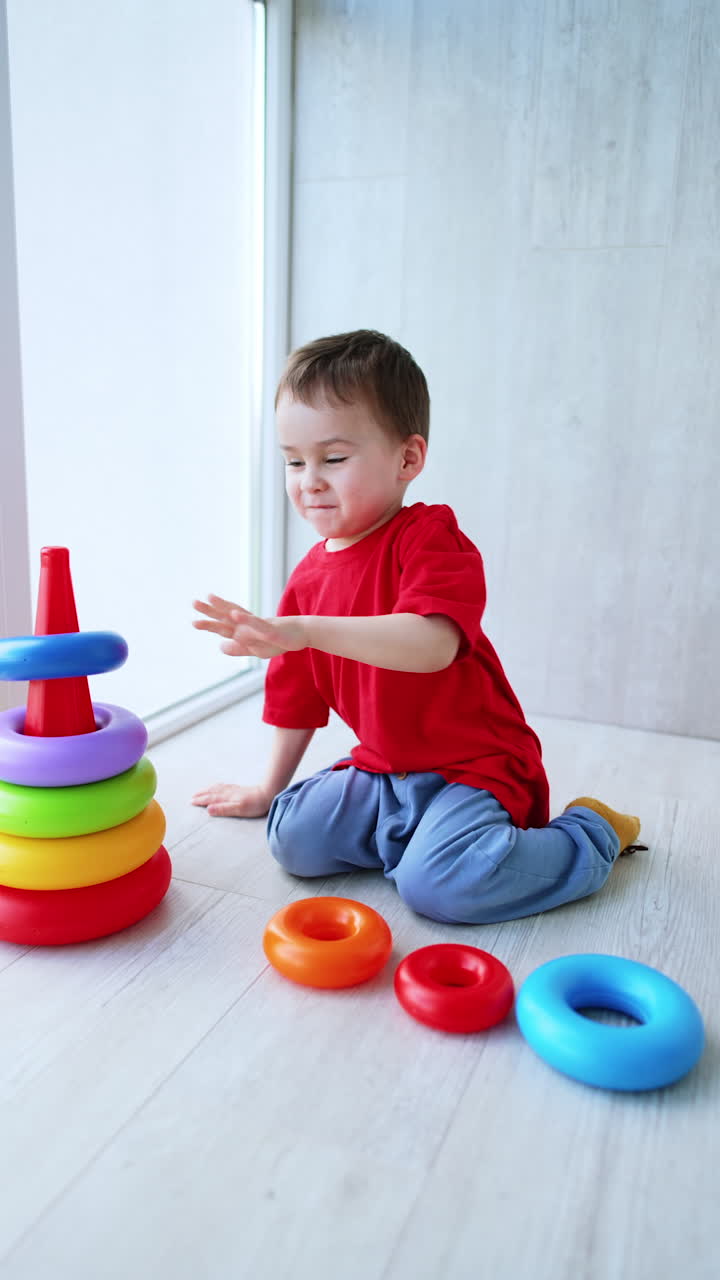 Adorable energetic kid sits on the floor playing with toy. Joyful toddler boy building a pyramid. Vertical video.