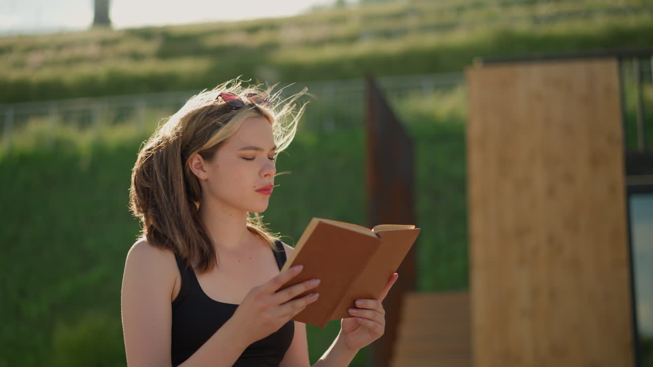 Woman seated, reading a book while wind gently blows her hair, sunlight illuminates her as she reads, with a lush greenery hill and wooden structure blurred in the background