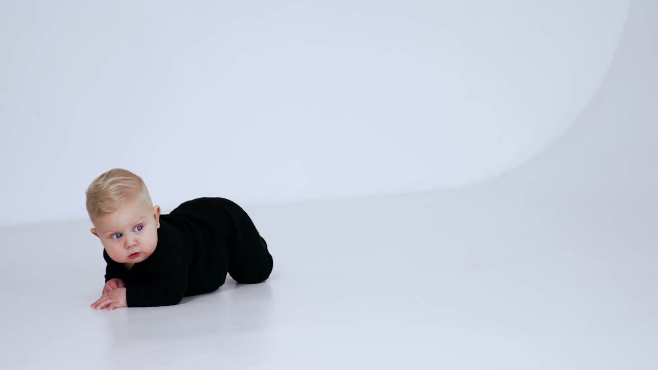 Little infant baby boy in black suit crawling by the floor. Adorable blond child at white backdrop.