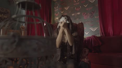 Fortune-teller in a plague doctor mask performing a spell in a chair in a living room decorated with butterfly wallpaper