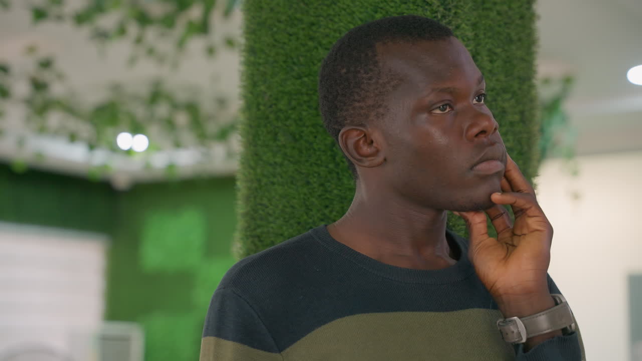 Contemplative urban portrait. Man contemplating solitude among lush indoor foliage under subdued light. Pensive individual seated near greenery carefully weighing thoughts during quiet moment indoors