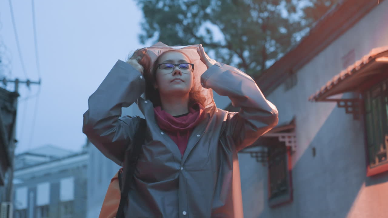 Donna asiatica che guarda in alto sotto un lampione, cappotto con cappuccio, borsa arancione, mani che aggiustano il cappuccio, bagliore morbido e caldo del lampione, espressione di meraviglia e risolutezza, color grading cinematografico, microavventura in un vicolo urbano.