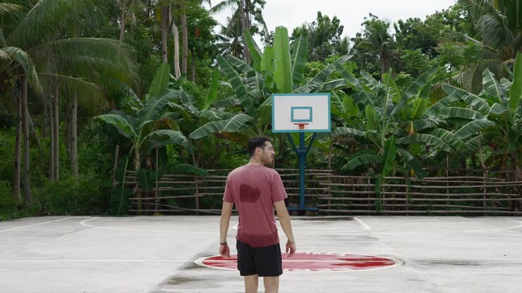Uomo in maglietta rossa che osserva un canestro da basket in un ambiente tropicale, di giorno, atmosfera sportiva casual.