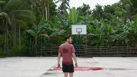 Man-in-red-shirt-gazing-at-basketball-hoop-in-tropical-setting,-daylight,-casual-sport-vibe