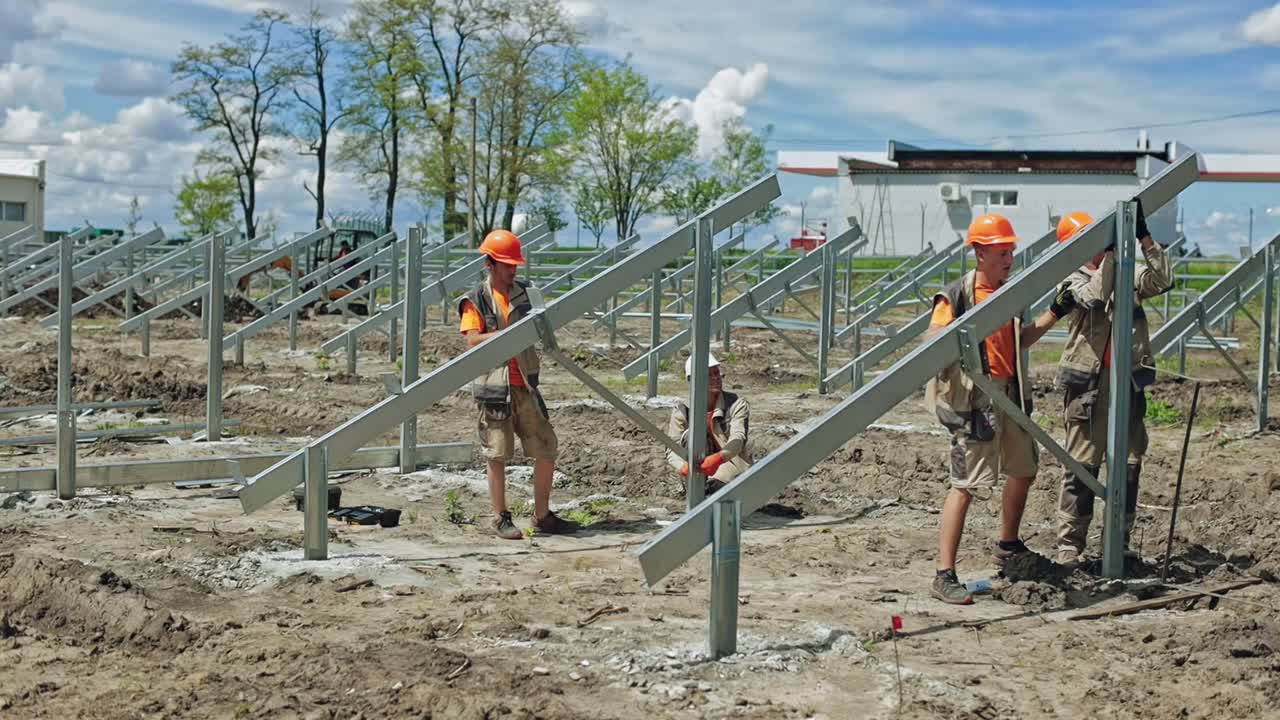 Engineers putting metal construction on the ground. Group of workers in hard hats build metal basis for solar panels on the field in summer day.