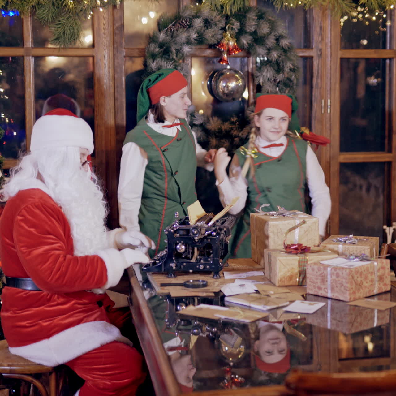 Joyful elves and Santa in festive costumes. Santa is busy at typewriter while two elves dancing inside Santa's residence. Christmas atmosphere.