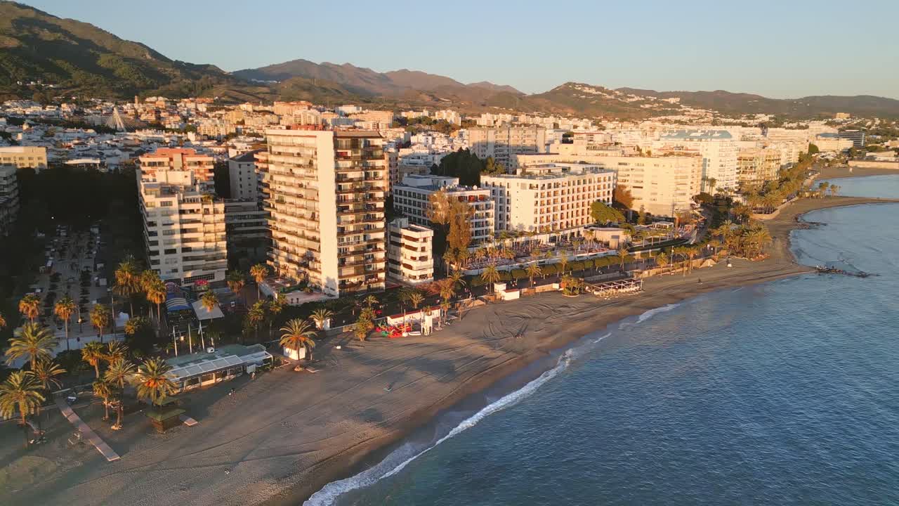Drone che vola verso una ripresa dall'alto sopra la spiaggia di Marbella, con vista dall'alto su lussuosi edifici costieri e sul lungomare illuminato dalla calda luce del tramonto, Andalusia, Spagna