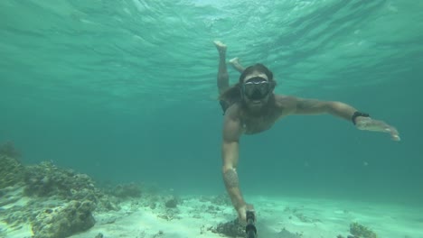 Un-Hombre-Joven,-En-Forma-Y-Fuerte-Con-Pelo-Largo-Y-Barba-Está-Nadando-Con-Gafas-De-Esnórquel-Se-Sumerge-Desde-La-Superficie-Hasta-El-Fondo-Del-Océano-Junto-A-La-Arena,-Rocas-Y-Arrecifes-Y-Vuelve-A-Nadar