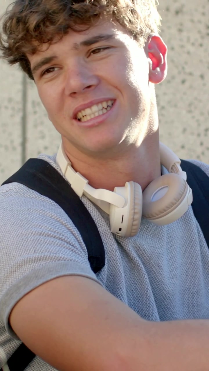 Vertical video: Smiling man responding to left person raising phone by campus wall, with headphones