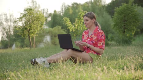 a middle-aged woman sits on the grass in the park. using a video link on a laptop, she conducts a conversation.