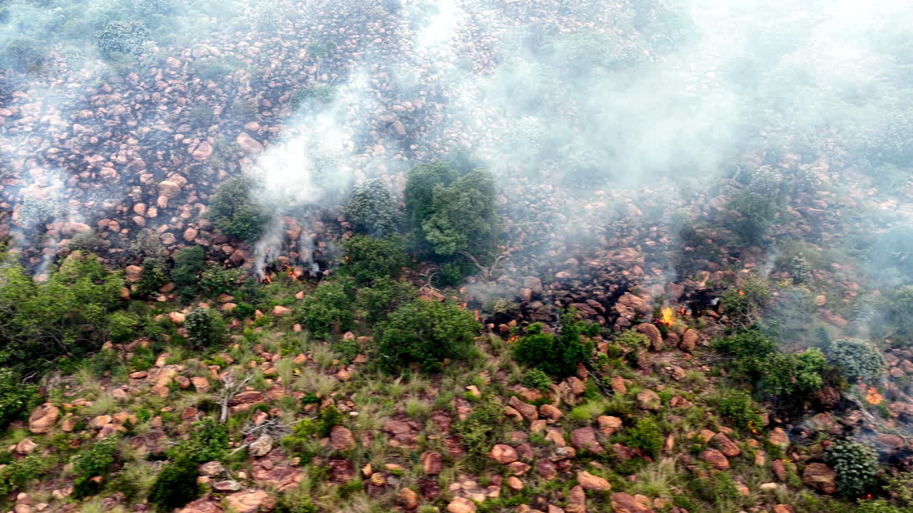 Il vento soffia contro il fronte di avanzamento di un incendio boschivo su un fianco di montagna roccioso, ripresa aerea.