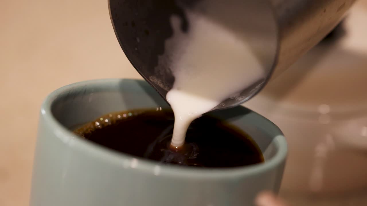 Milk is poured into a steaming cup of coffee, creating swirling patterns. Warm lighting enhances the cozy atmosphere