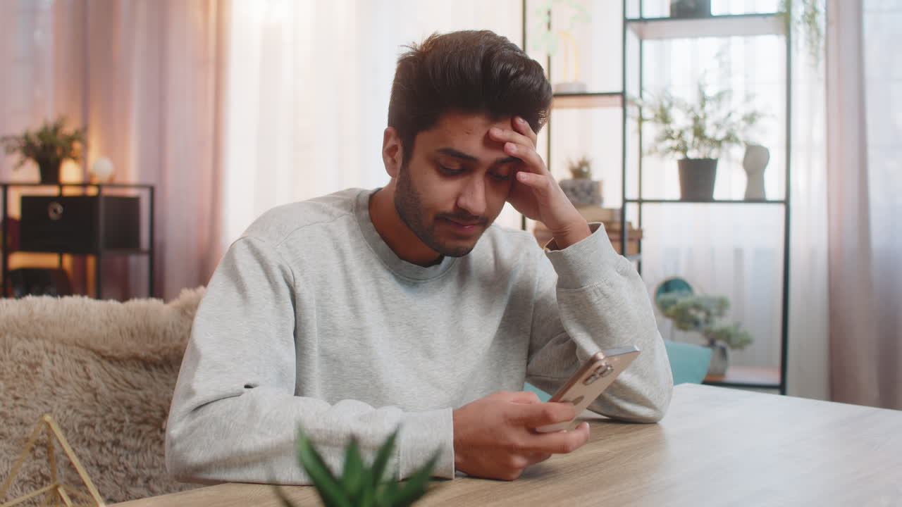 Young indian man texting on smartphone while resting head on hand at table in living room at home